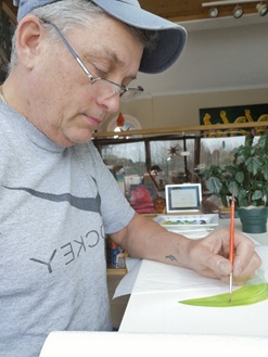 Man in a cap and glasses painting at a table.