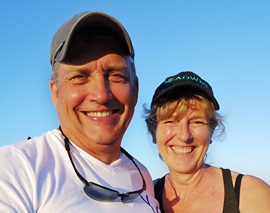 Two smiling adults wearing caps and sunglasses outdoors during sunset.